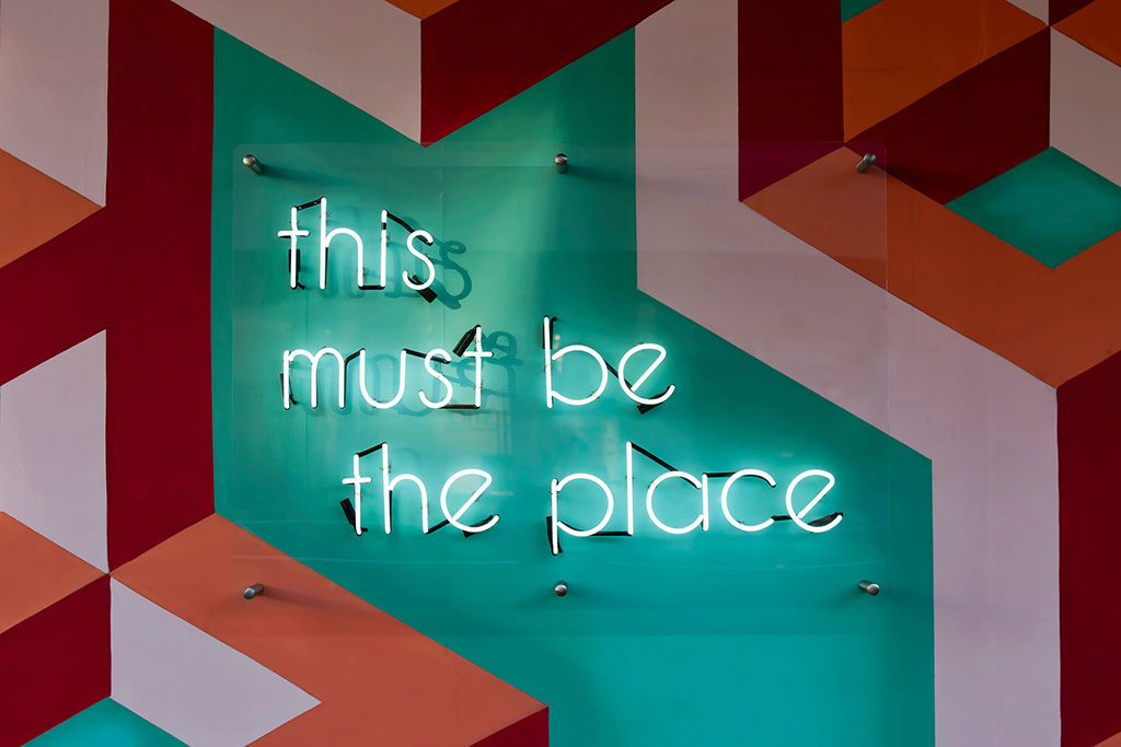 A photograph of a painted wall, which has a neon sign which says "this must be the place"
